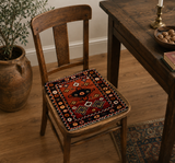 A rustic wooden dining chair in a cozy room setting, featuring a colorful geometric kilim seat cushion with red, orange, and black patterns.