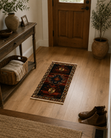 A long runner rug with a traditional geometric pattern in dark red, black, and beige is styled on a light wood entryway floor. The scene includes a wooden front door, a console table with a basket and vase, potted plants, and a pair of brown boots on the floor.